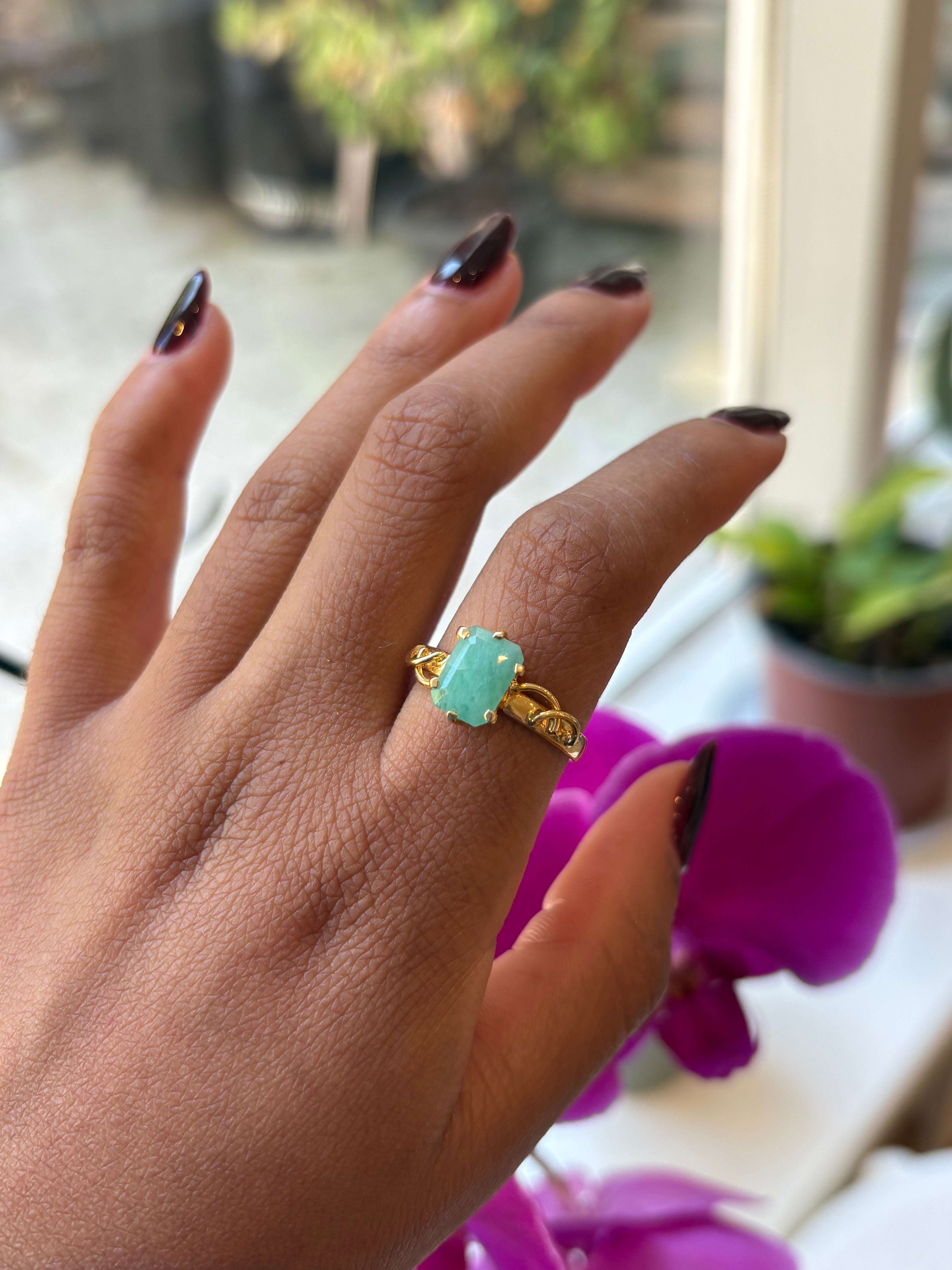 A gold ring with a light green rectangular stone worn on a person's ring finger, shown against a blurred background with a purple orchid flower and a window.